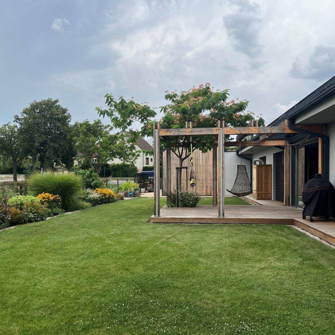 Weitläufiger Garten mit Holzterrasse, Pergola und blühenden Beeten am Haus.