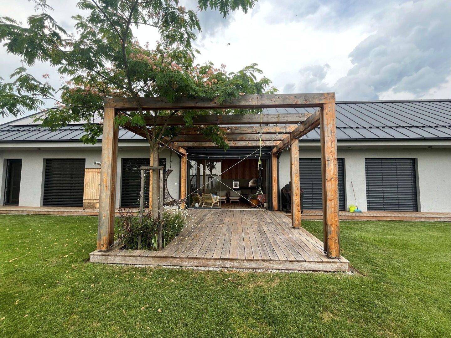 Holzterrasse mit Pergola vor dem modernen Haus mit großen Fenstern und Rollläden.
