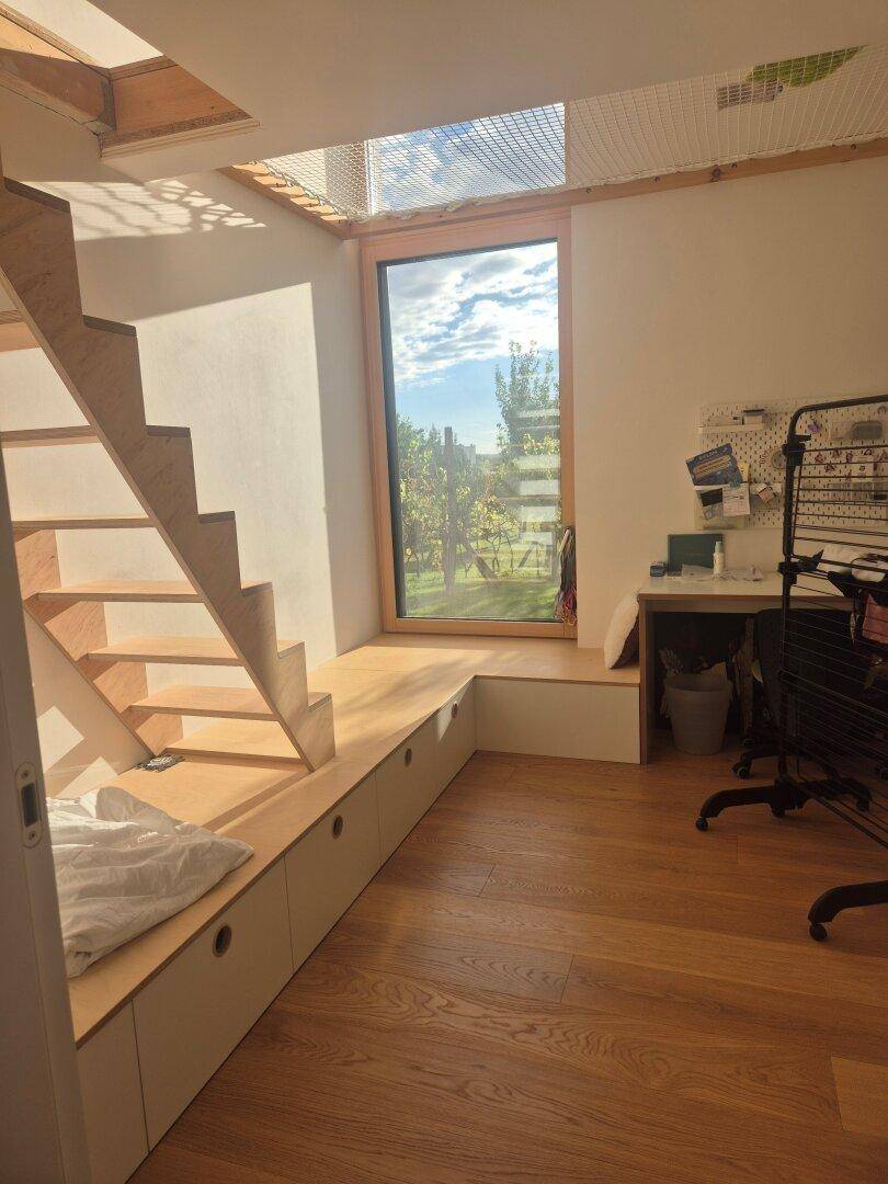 Helles Zimmer mit Holztreppe, integrierter Sitzbank und großem Fenster mit Blick ins Grüne.