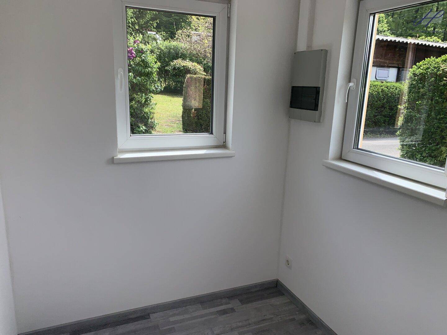 Heller, leerer Raum mit zwei Fenstern und Blick ins Grüne, ausgestattet mit Laminatboden.
