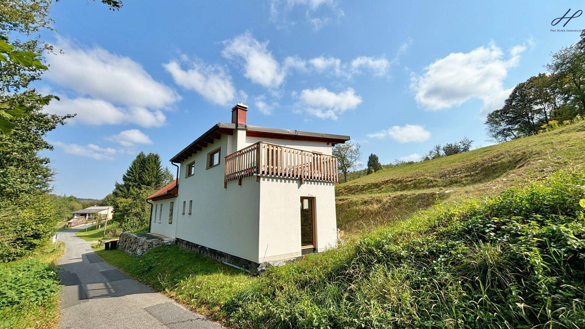 Seitenansicht des weißen Hauses mit Holzbalkon und Blick auf die umliegende grüne Hügellandschaft.