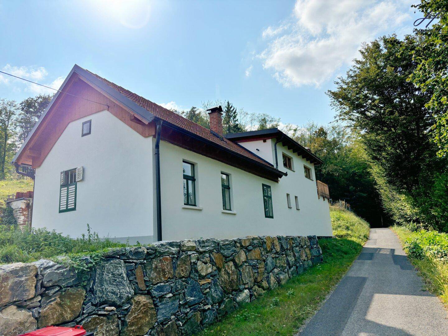 Seitenansicht des weißen Hauses mit Steinmauer und grünen Fensterläden, umgeben von üppiger Natur.