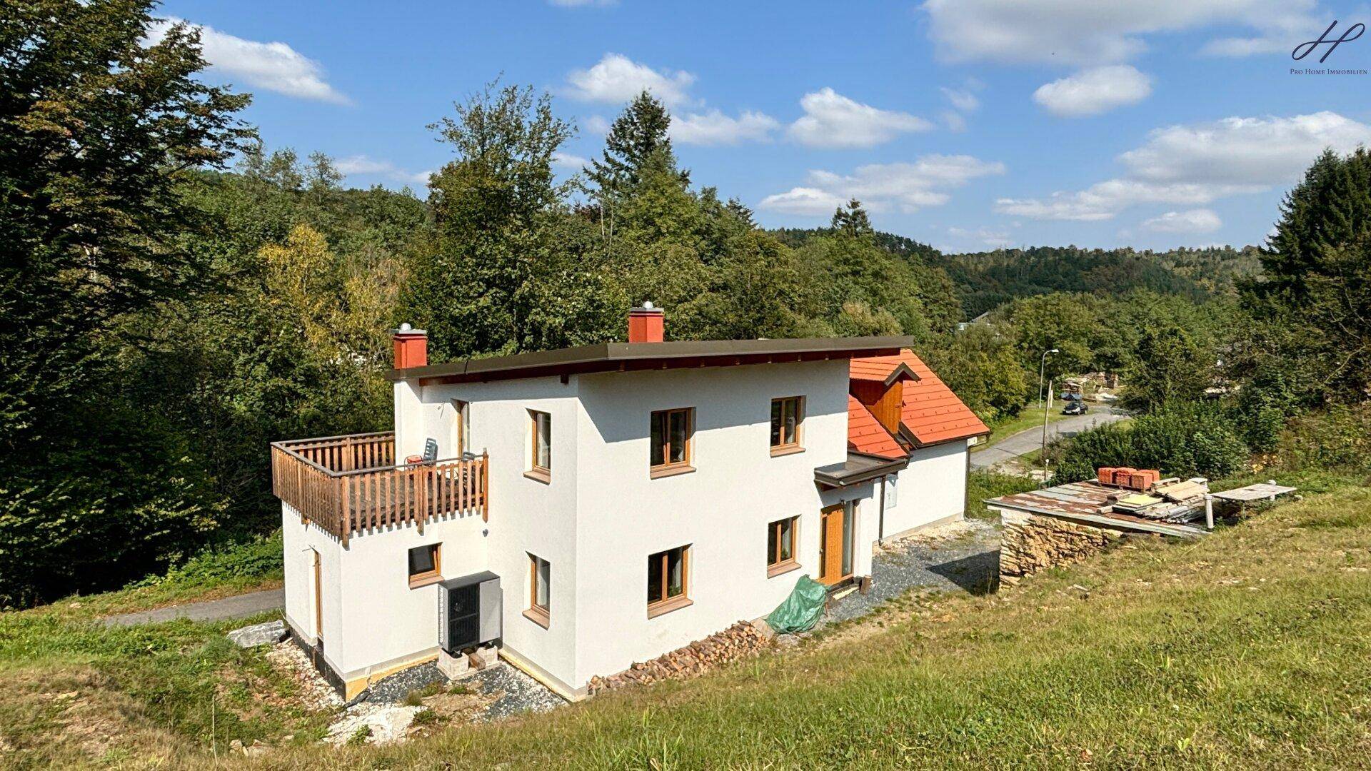 Weißes Einfamilienhaus mit Balkon und rotem Ziegeldach, umgeben von grüner Natur unter blauem Himmel.