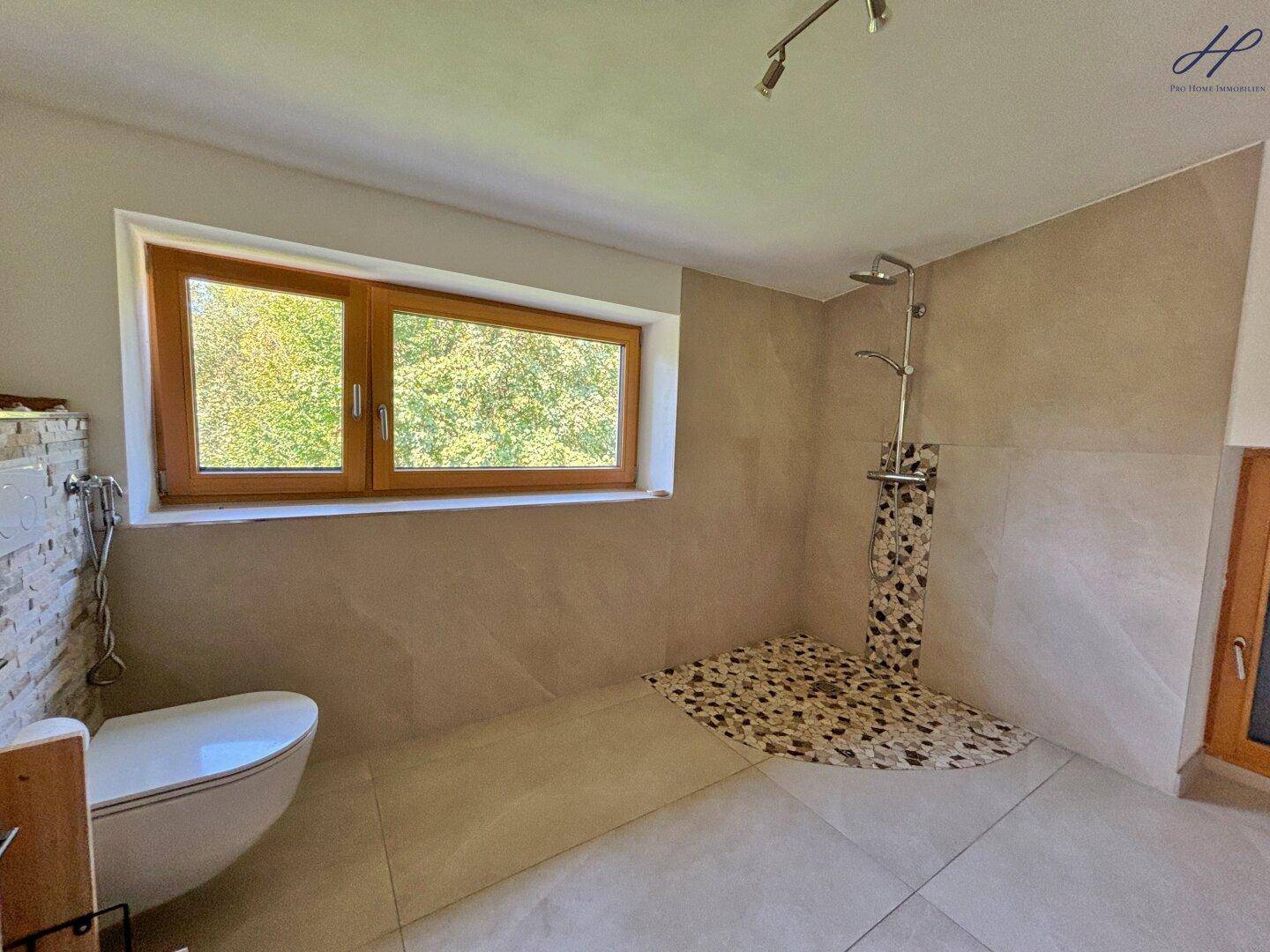 Geräumiges Badezimmer mit begehbarer Dusche, Wand-WC und Fenster mit Blick ins Grüne.