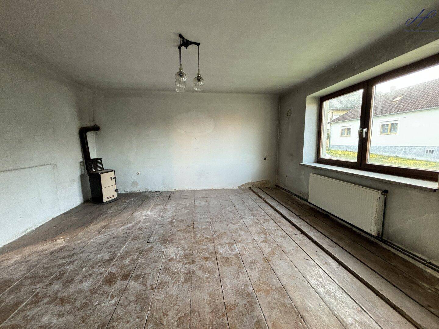 Leerer Raum mit altem Dielenboden, einem Fenster und einem traditionellen Ofen.