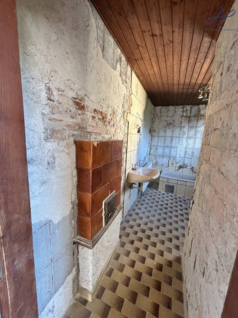 Badezimmer im Renovierungszustand mit alten Fliesen, Waschbecken und einem Kachelofen.