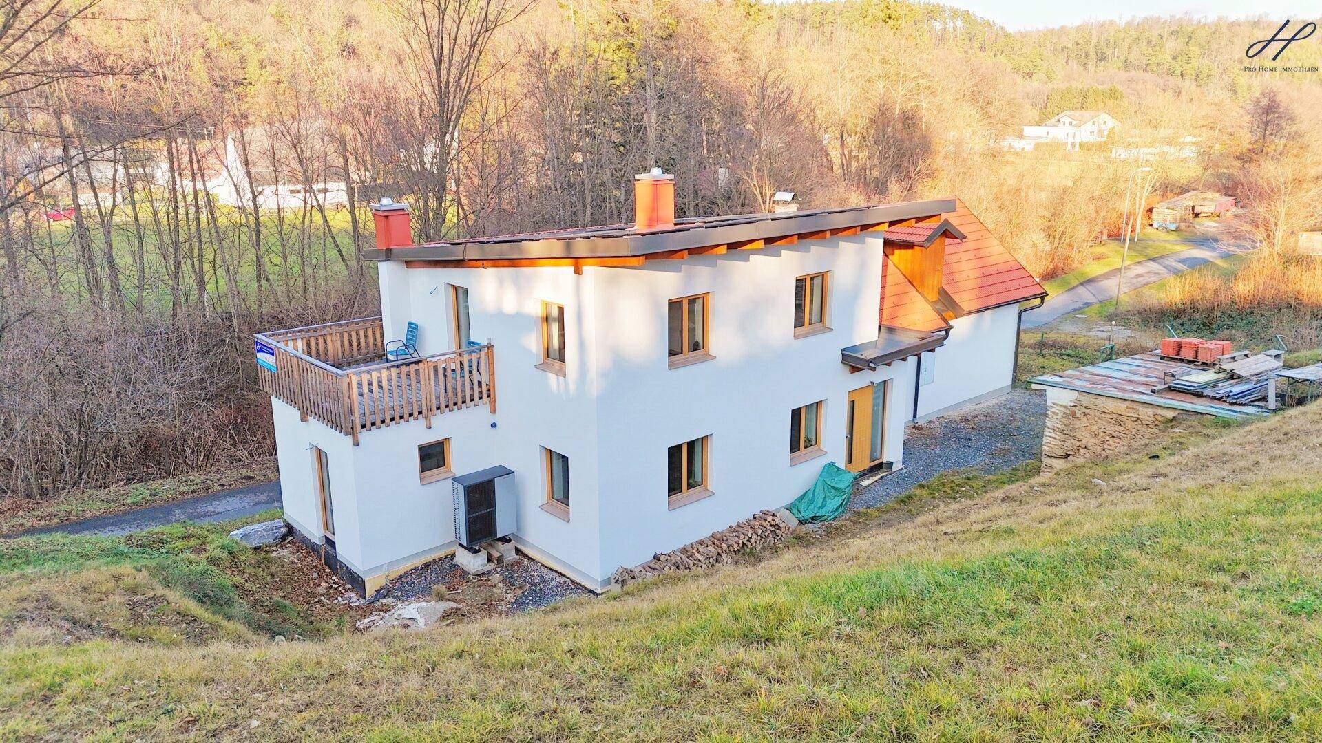 Weißes Einfamilienhaus mit Balkon und rotem Ziegeldach, umgeben von grüner Natur im Sonnenlicht.