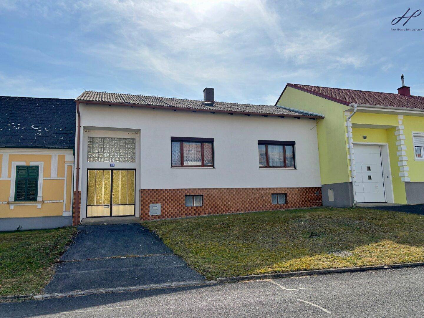 Vorderansicht des Hauses mit Zufahrt und angrenzenden Gebäuden an einem sonnigen Tag.