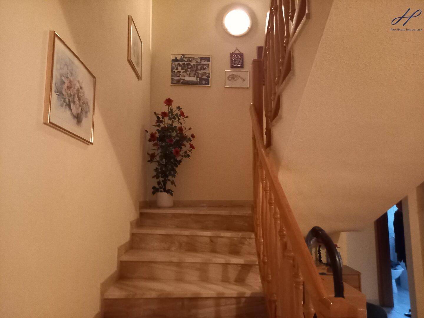 Treppenhaus mit Holzgeländer und Bildern an der Wand, beleuchtet von einer runden Deckenleuchte.