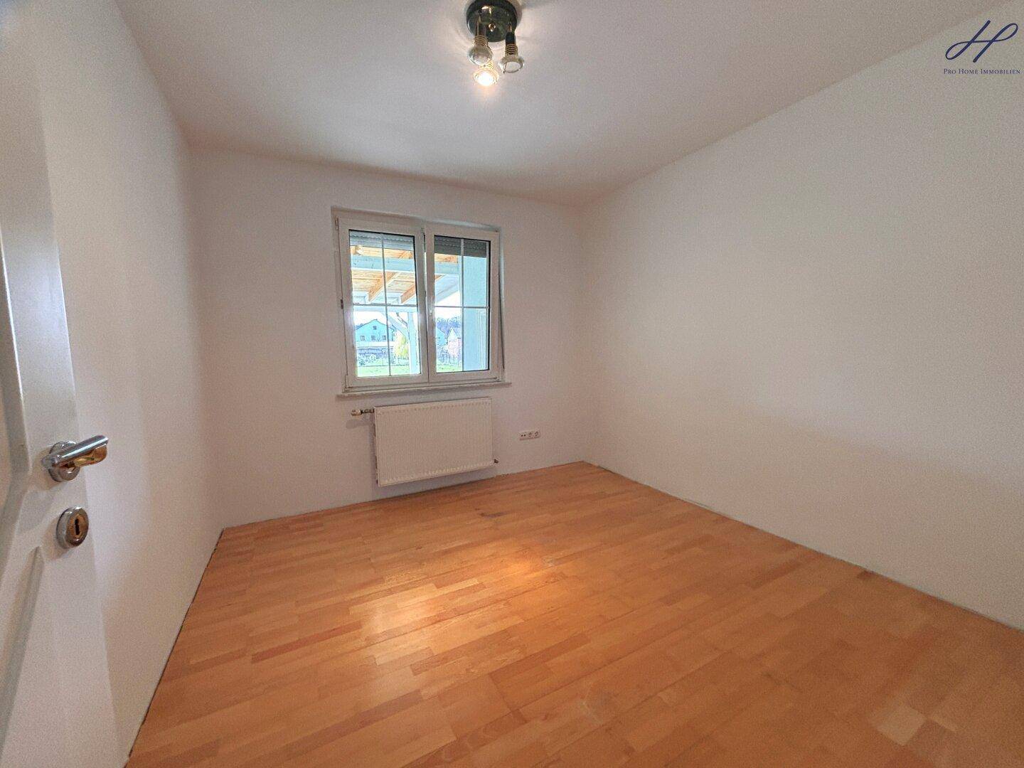 Helles Zimmer mit Holzparkettboden und Fenster, unmöbliert bis auf einen Heizkörper.