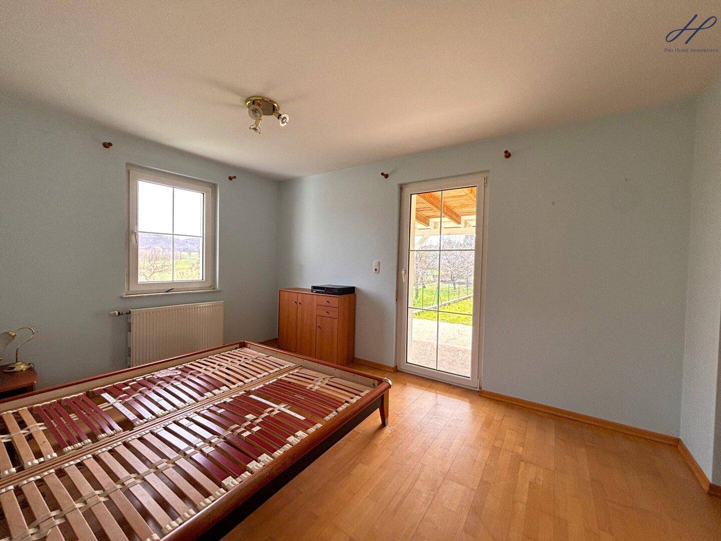 Schlafzimmer mit Holzparkettboden, Fenster und Zugang zur überdachten Terrasse.