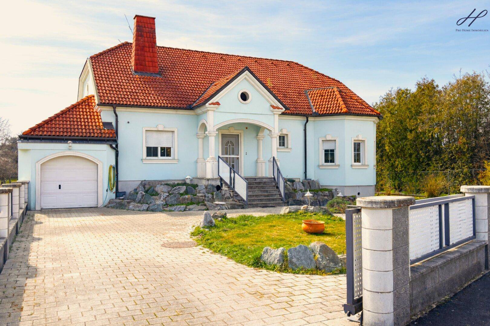 Frontansicht eines gepflegten Einfamilienhauses mit hellblauer Fassade, rotem Ziegeldach und gepflasterter Auffahrt.