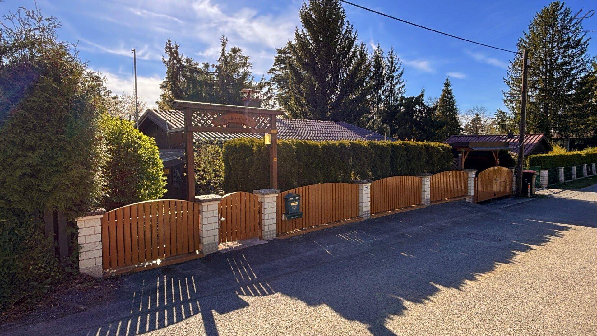 Eingangsbereich des Grundstücks mit Holzzaun, Hecke und Carport im Hintergrund.