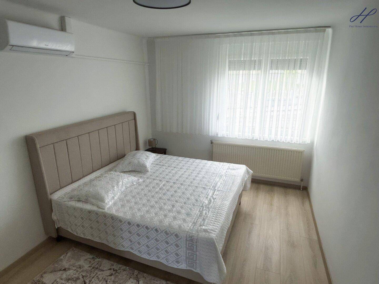 Ruhiges Schlafzimmer mit Doppelbett, hellem Laminatboden, großem Fenster und Klimaanlage.