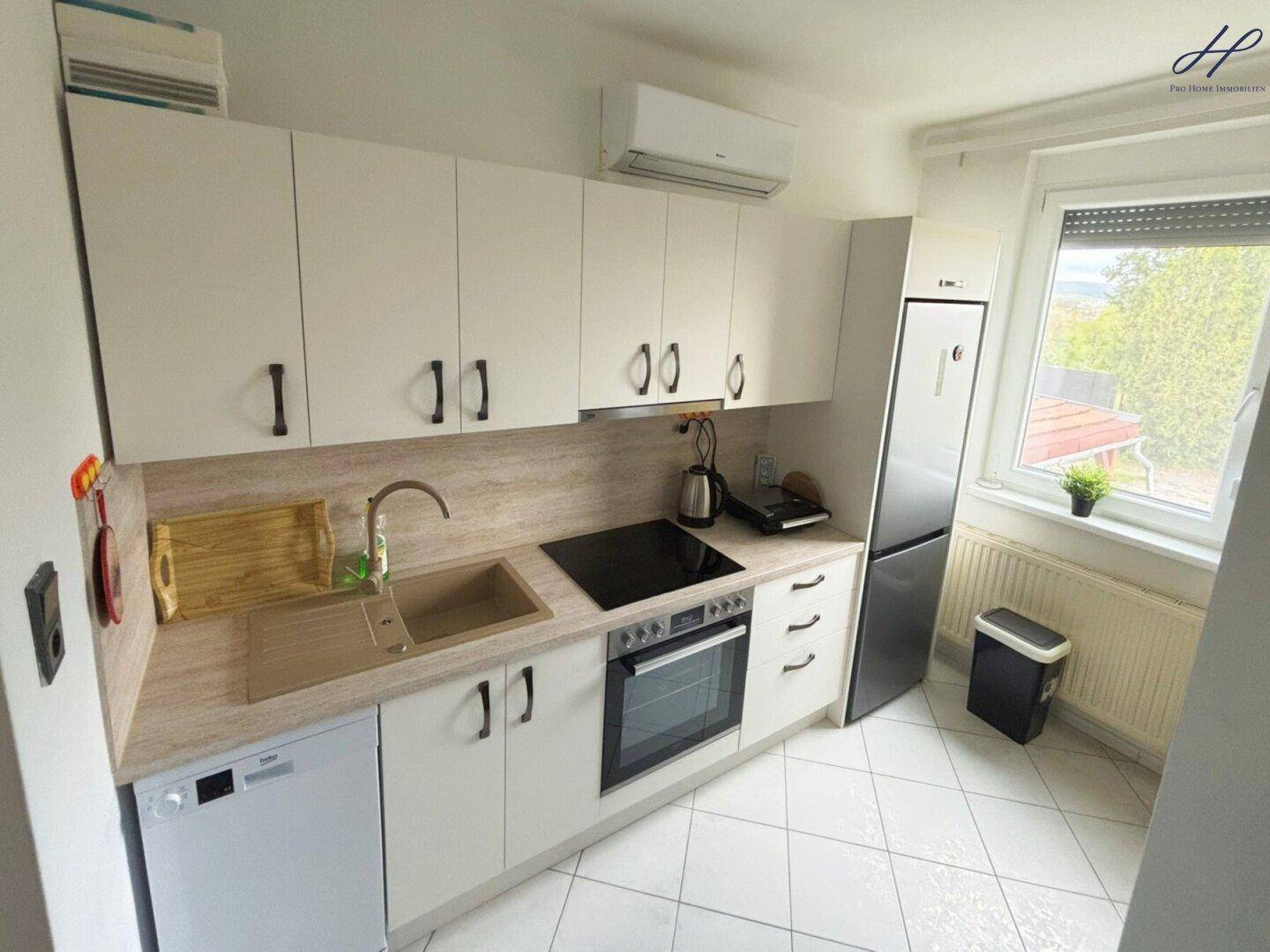 Funktionale Küche mit weißen Schränken, Induktionskochfeld, Backofen und einem Fenster mit Blick ins Grüne.