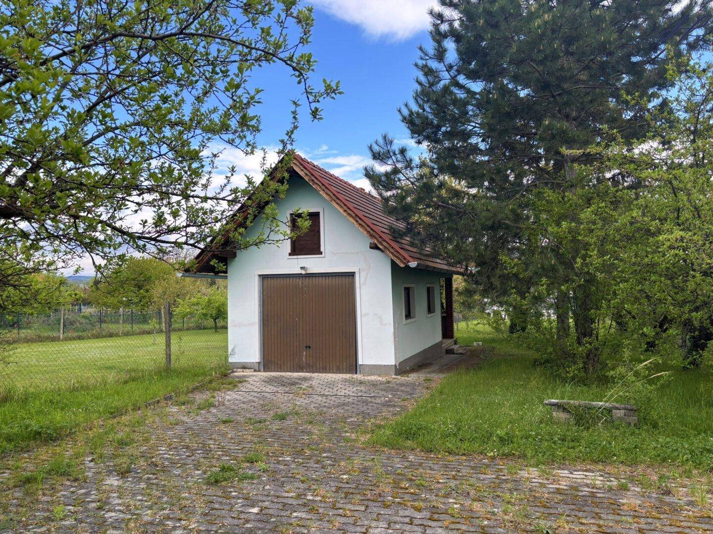 Freistehende Garage mit Satteldach und braunem Tor, umgeben von viel Grün und Bäumen.