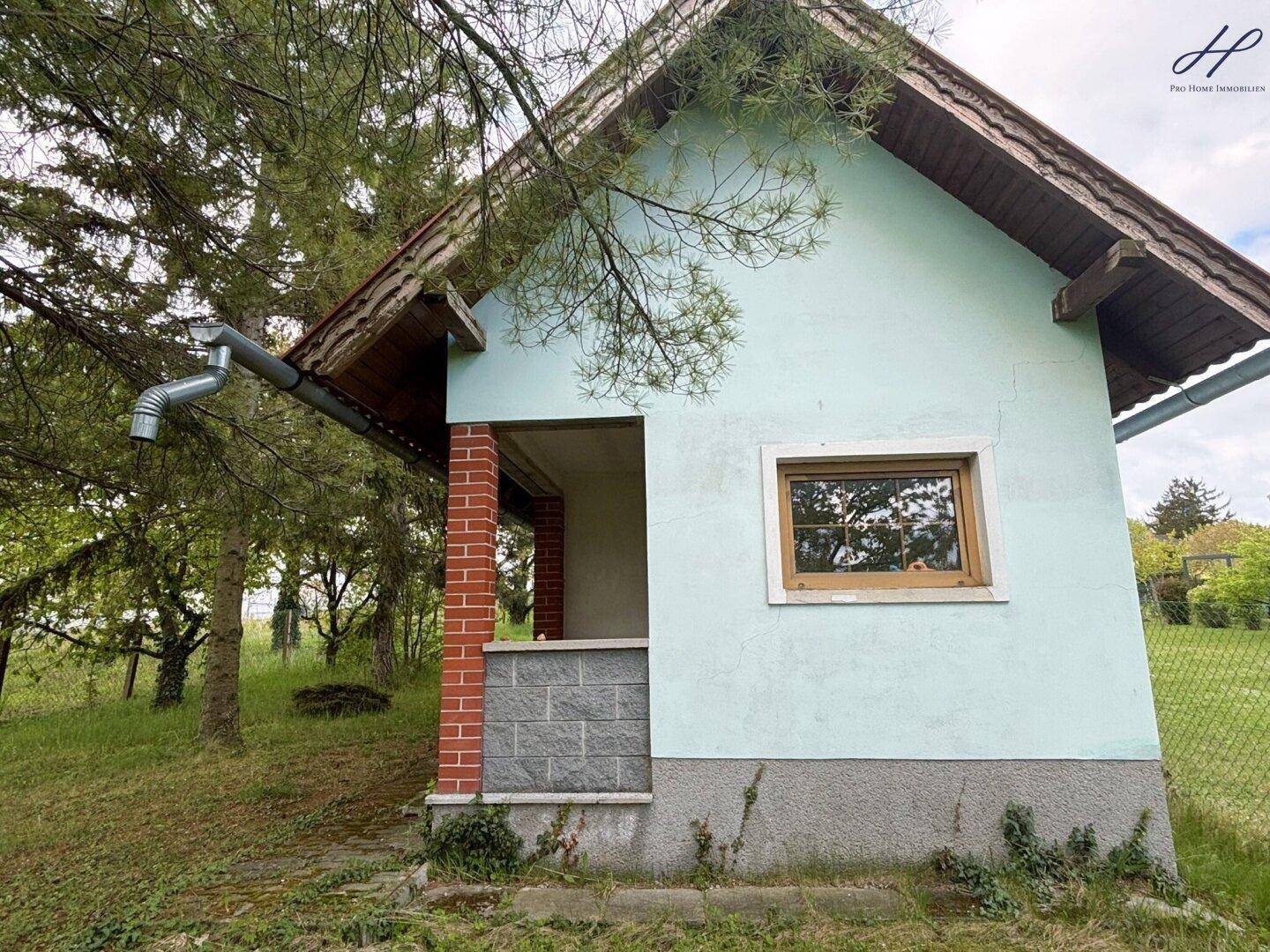 Kleines Nebengebäude mit hellgrüner Fassade und Ziegelsockel, umgeben von Bäumen und Natur.