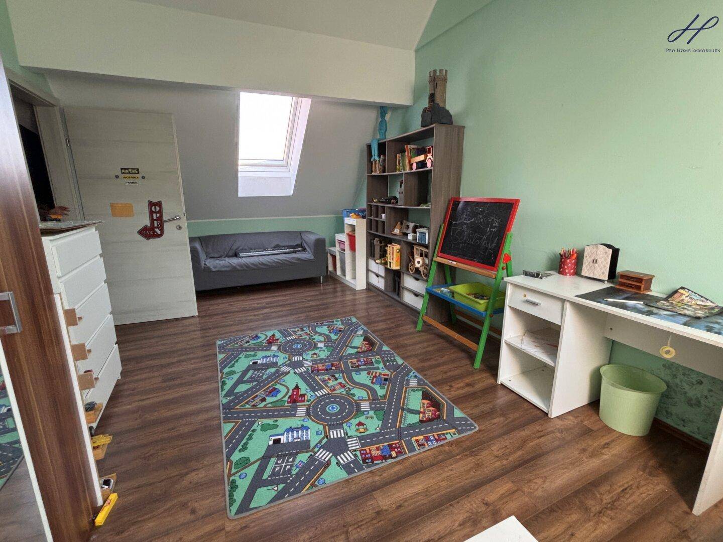 Geräumiges Kinderzimmer mit Holzboden, Spielteppich und einem gemütlichen Sofa unter dem Dachfenster.