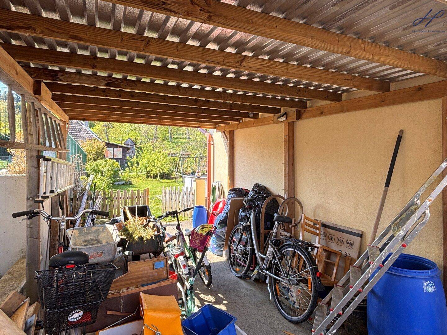 Überdachter Abstellbereich mit Fahrrädern, Leitern und verschiedenen Gegenständen, umgeben von Natur.