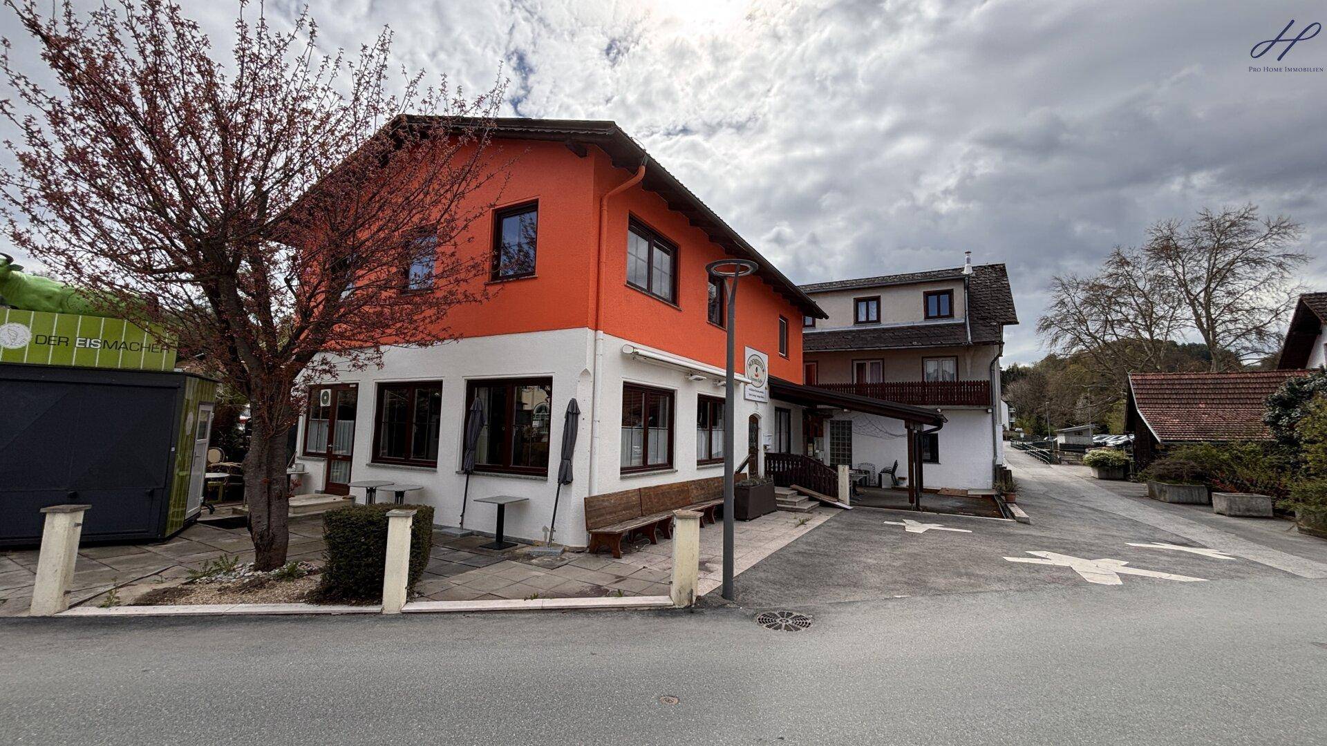 Seitenansicht des Gebäudes mit orangefarbener Fassade und einem blühenden Baum im Vordergrund, an einer Straße gelegen.