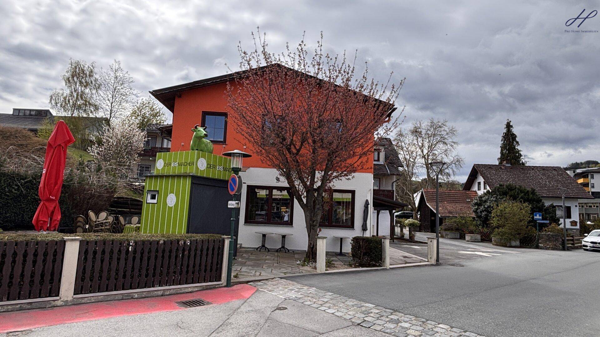 Gebäude mit orangefarbener Fassade und einem blühenden Baum, umgeben von einer Straße und weiteren Häusern im Hintergrund.