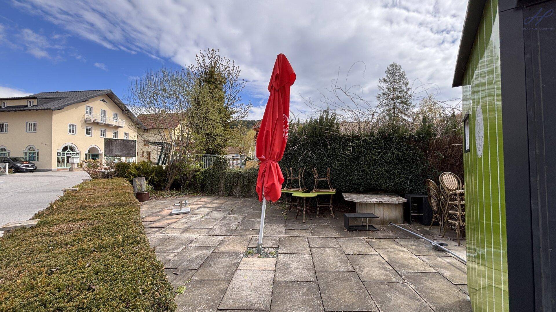 Große gepflasterte Terrasse mit einem roten Sonnenschirm, Gartenmöbeln und einer Hecke im Hintergrund.