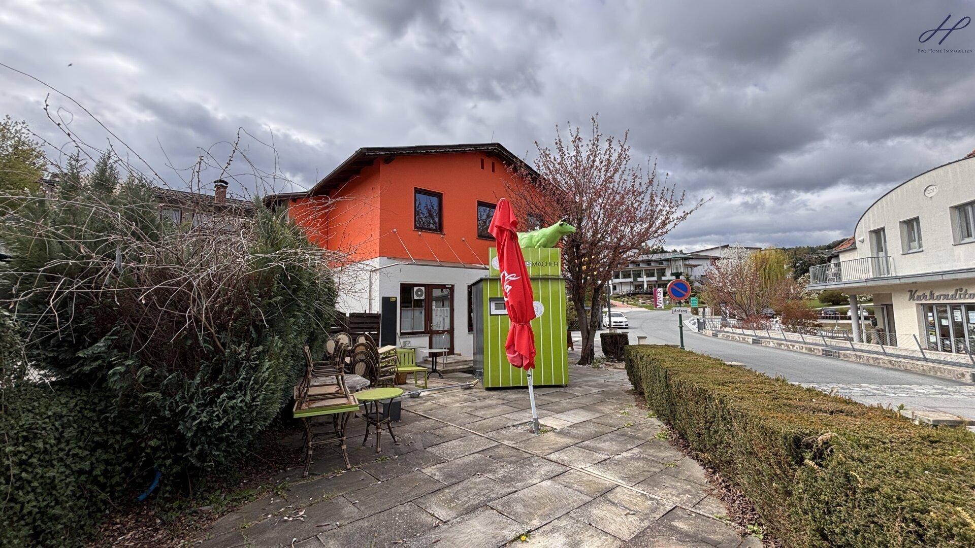 Gepflasterte Terrasse mit rotem Sonnenschirm und Gartenmöbeln vor einem Gebäude mit orangefarbener Fassade.