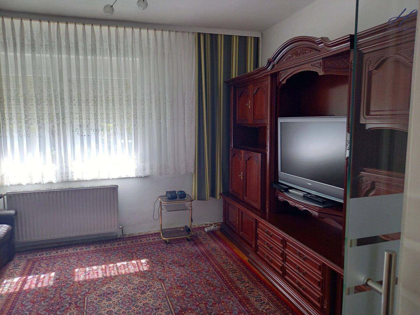 Wohnzimmer mit großem Fenster, Teppichboden und klassischem Holzschrank.