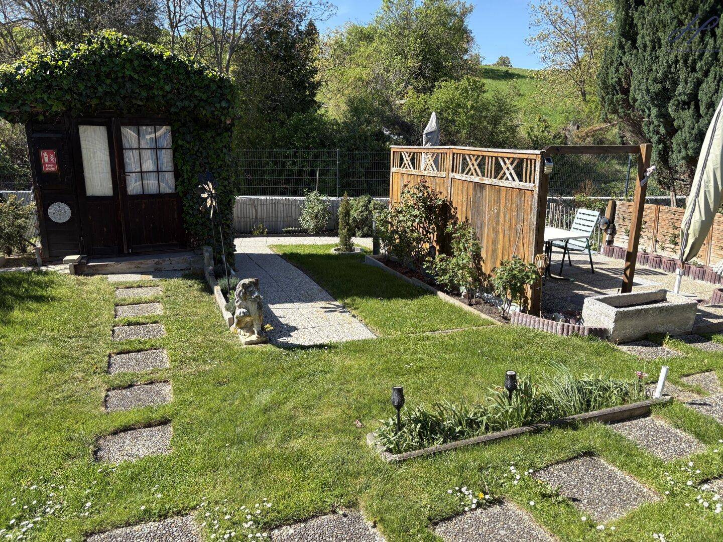 Grüner Gartenbereich mit Gartenhaus, gepflasterten Wegen und einer kleinen Terrasse im Hintergrund.