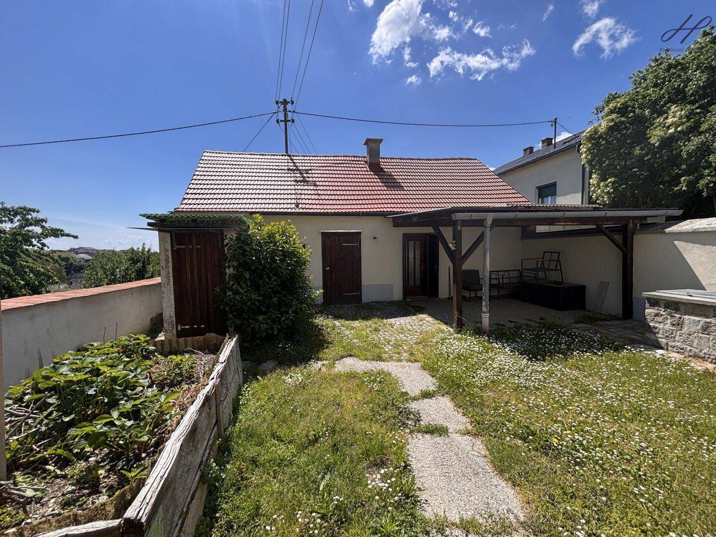 Ländliches Haus mit rotem Ziegeldach, grünem Garten und überdachtem Bereich unter blauem Himmel.