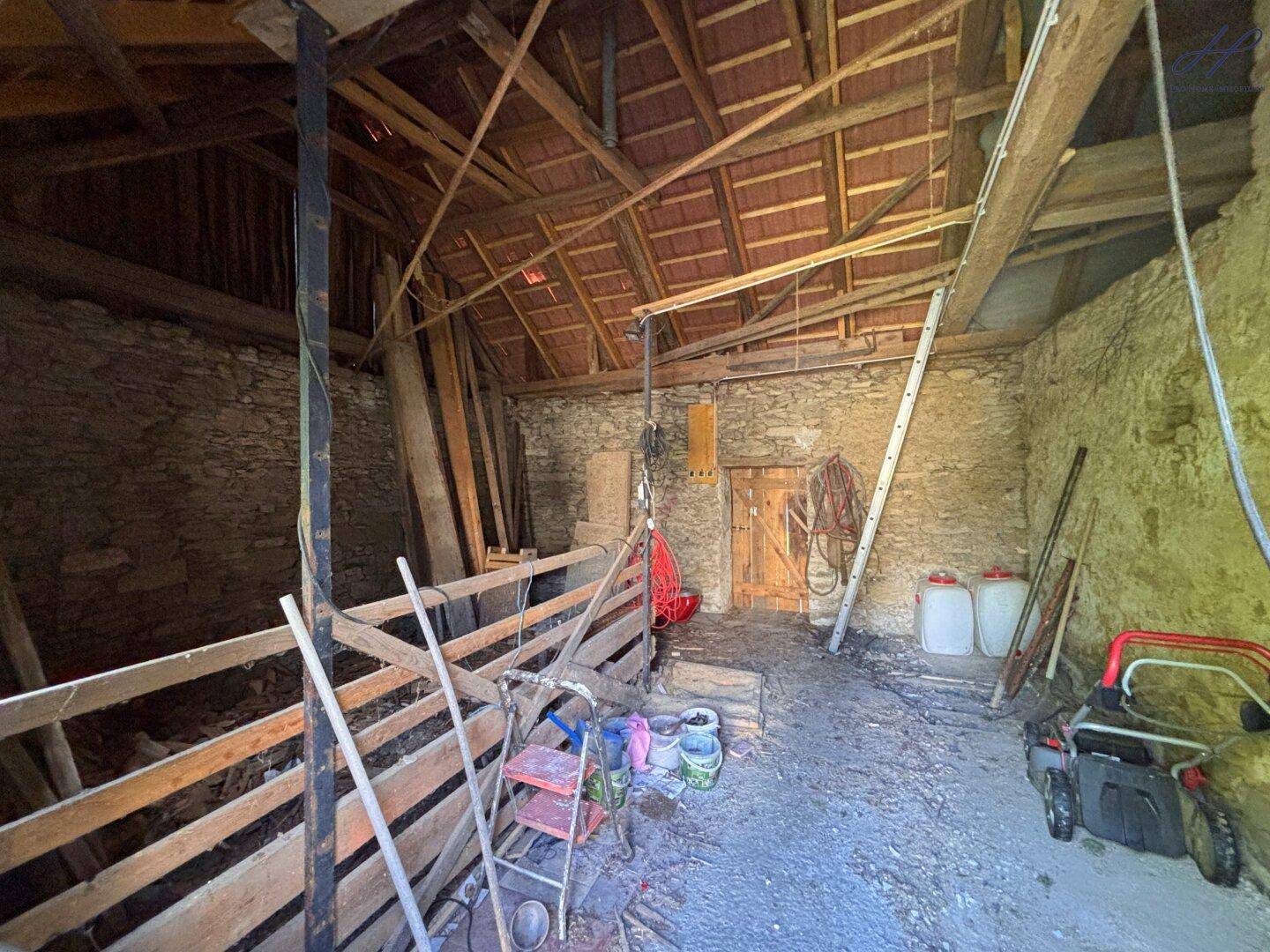 Geräumiger Innenraum eines Nebengebäudes mit Holzdachstuhl, Steinwänden und Lagerflächen.