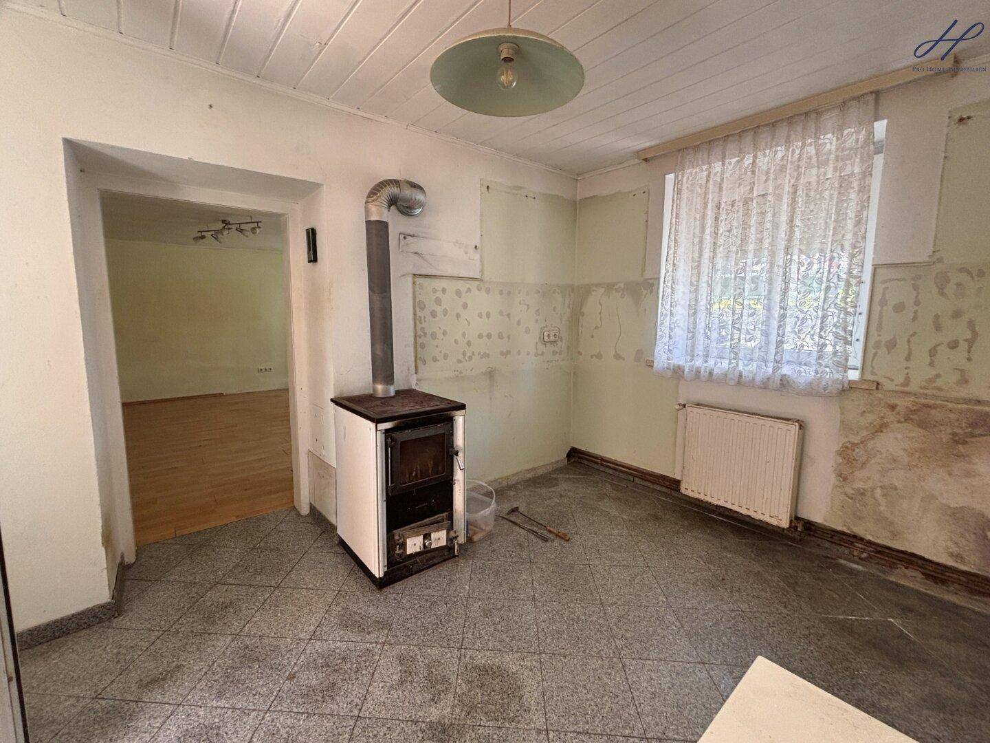 Renovierungsbedürftige Küche mit Kachelofen, Fliesenboden und Blick in einen weiteren Raum.