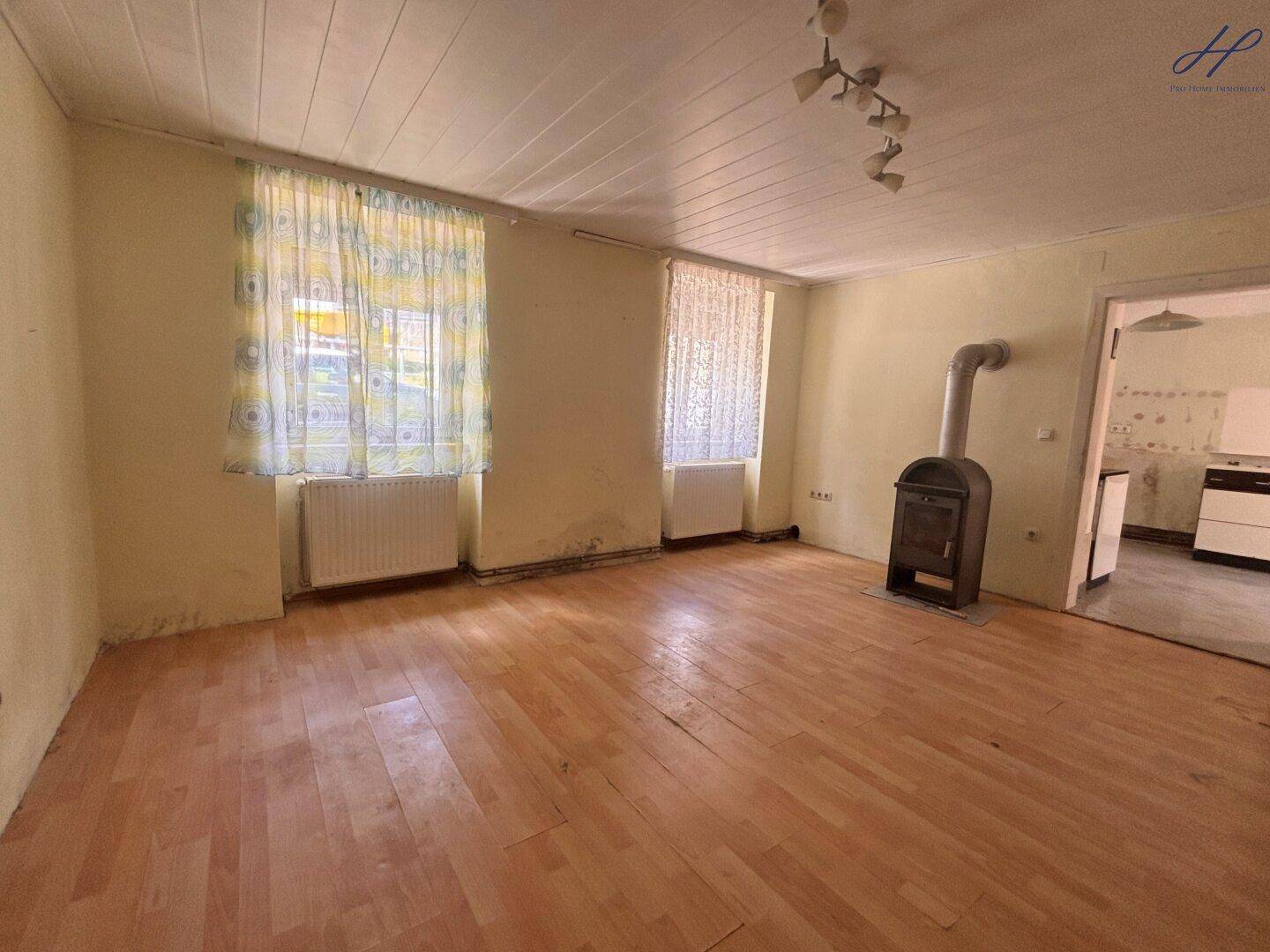 Geräumiges Wohnzimmer mit Holzboden, zwei Fenstern und einem Kachelofen.