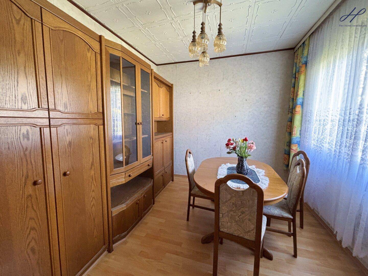 Esszimmer mit Holzschrank, Esstisch und Stühlen, sowie einem Fenster mit Vorhängen.