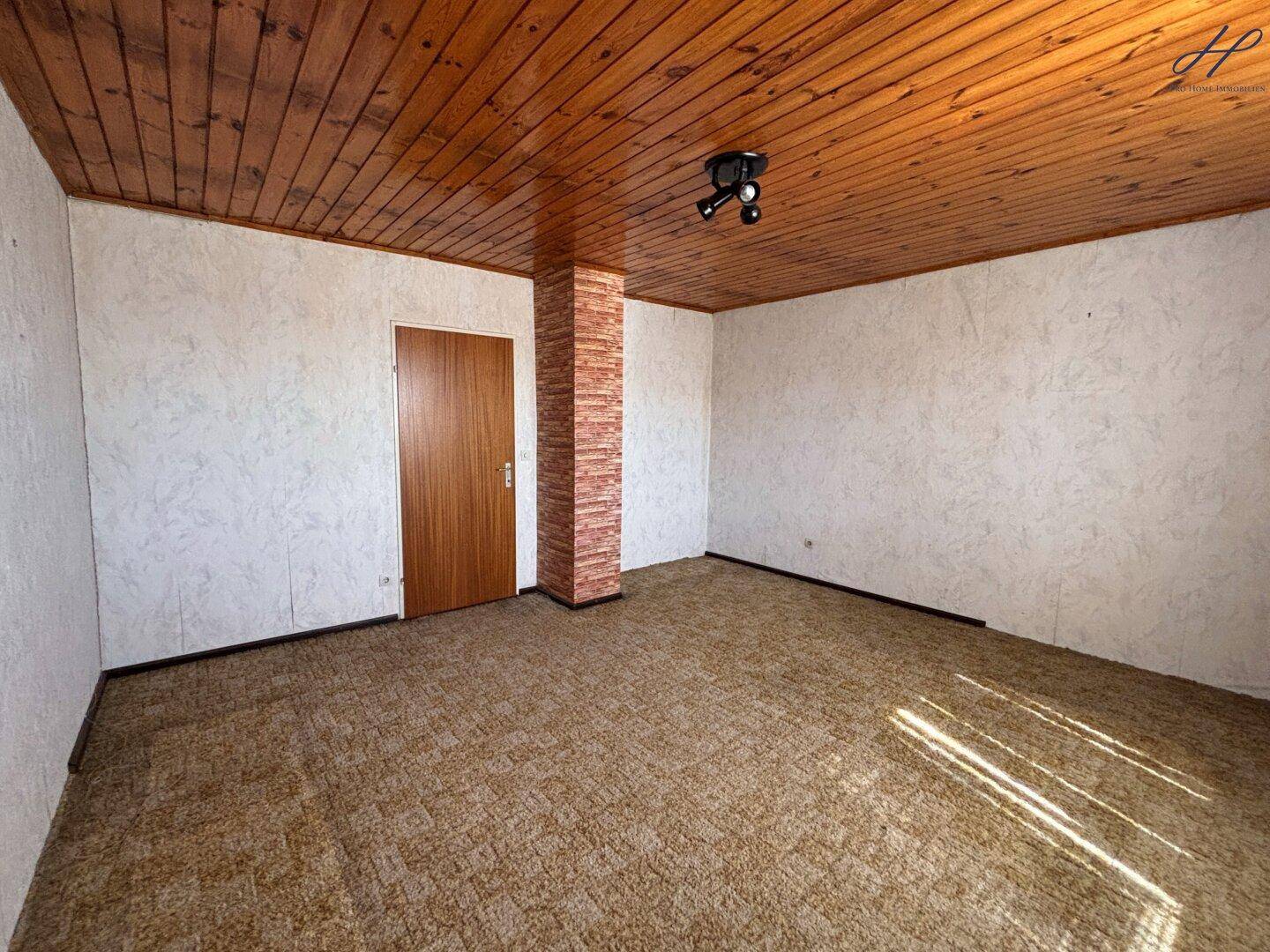 Geräumiges Wohnzimmer mit Teppichboden, Holzdecke und einer Ziersäule aus Backstein.