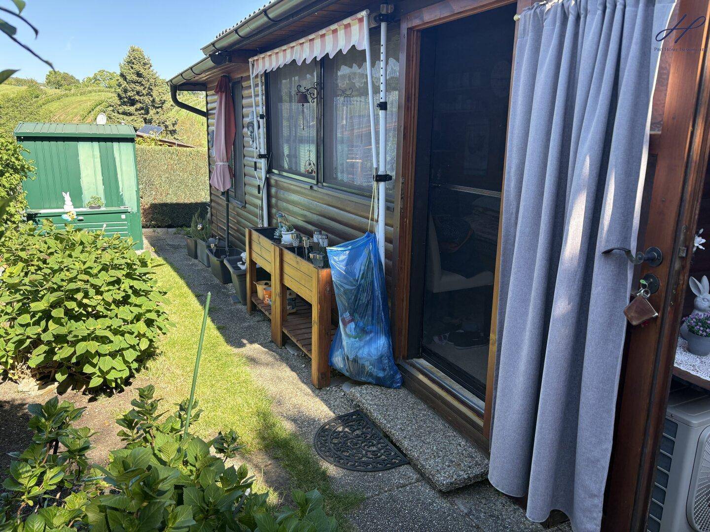 Seitlicher Gartenbereich mit Rasenfläche, Sträuchern und Zugang zur Holzhütte, umgeben von Natur.