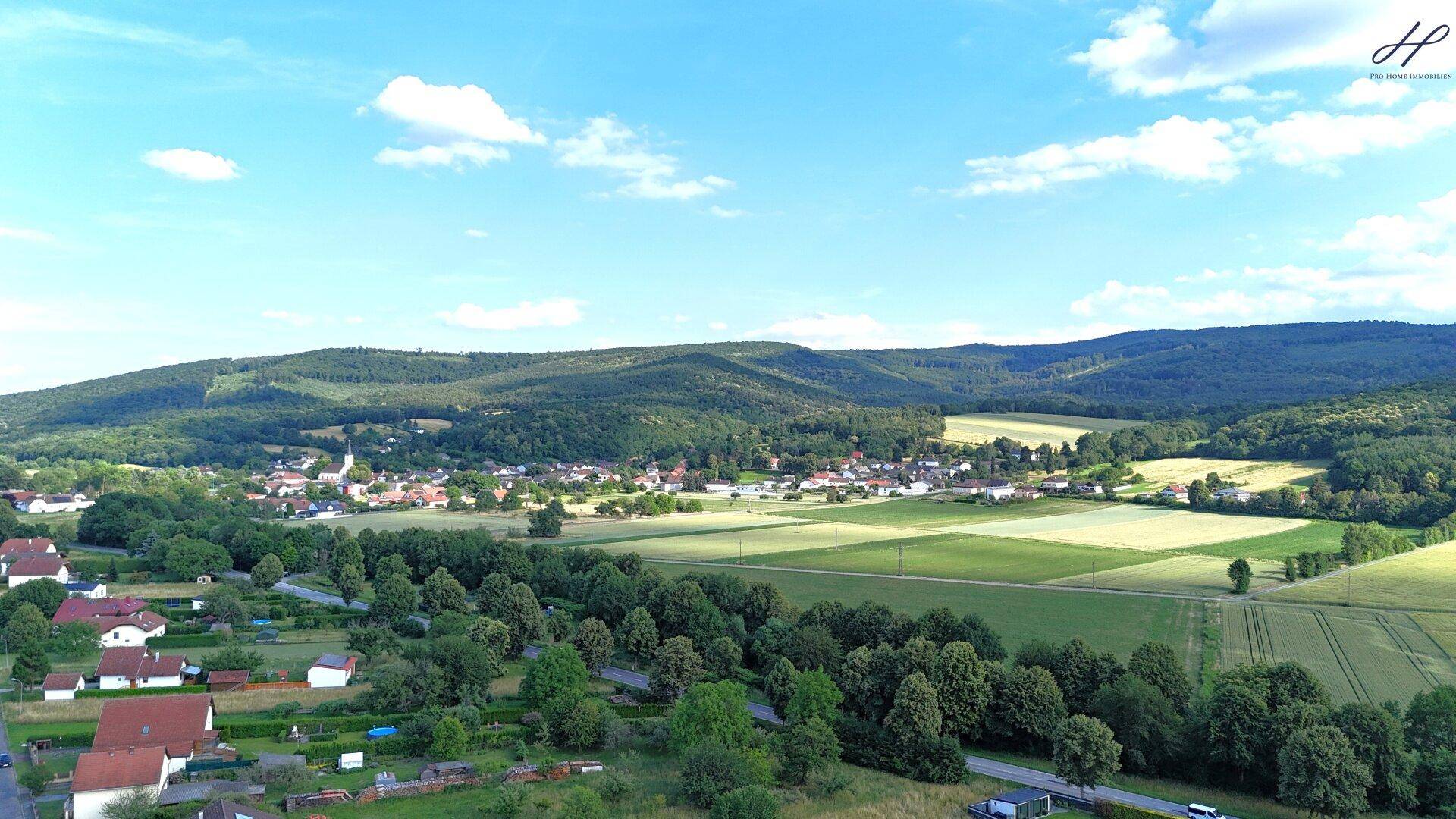Luftaufnahme eines Dorfes eingebettet in eine hügelige, grüne Landschaft.