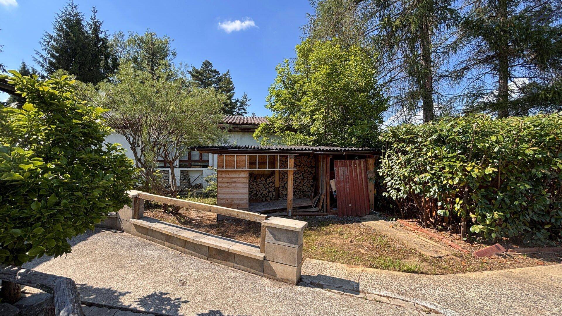Seitenansicht des Grundstücks mit einem Holzschuppen und dichter Bepflanzung im Hintergrund.