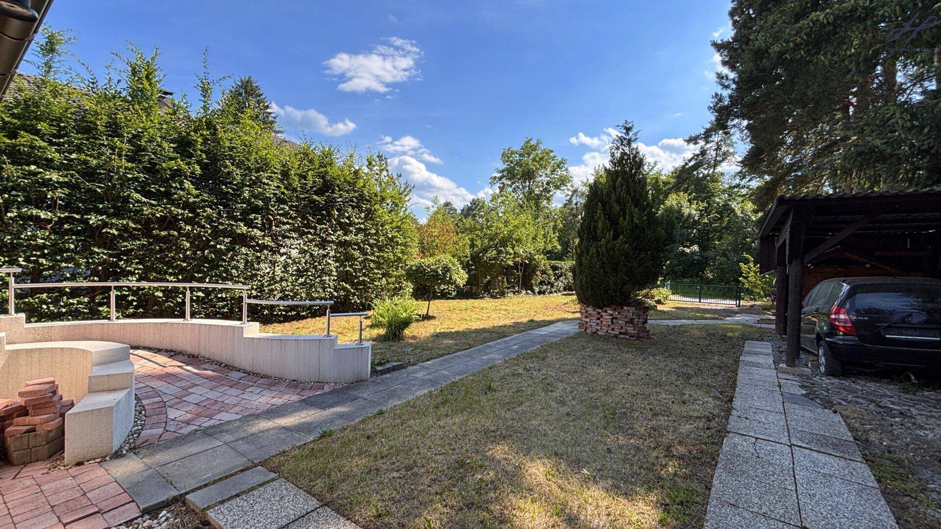 Blick vom Terrassenbereich in den Garten mit einem Weg, Rasenfläche und einem Carport.