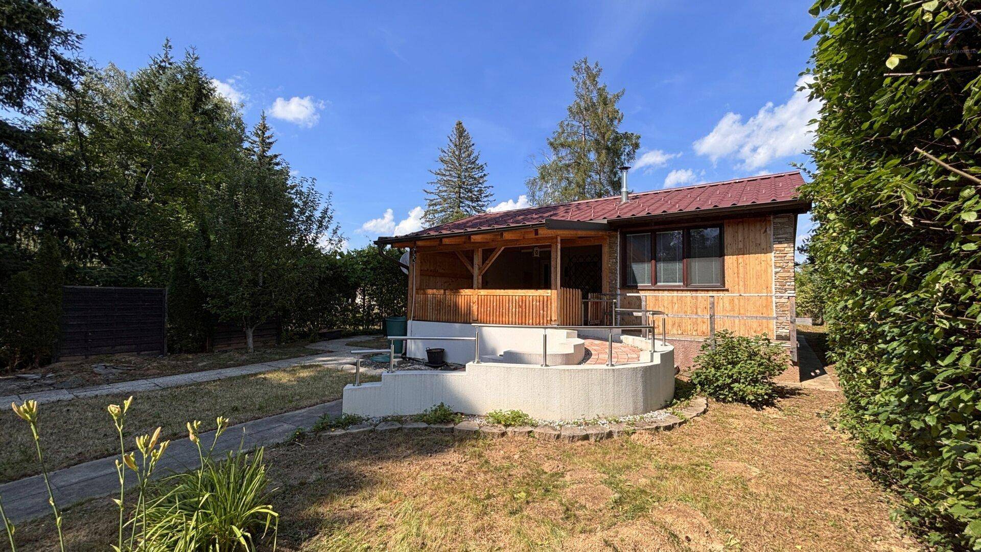 Seitenansicht des Hauses mit der überdachten Veranda, umgeben von Garten und Bäumen.
