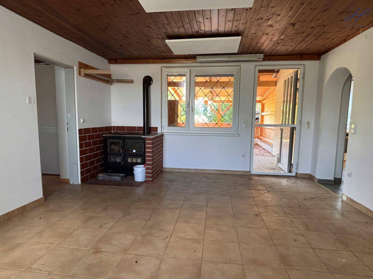 Gemütliches Wohnzimmer mit Fliesenboden, Holzdecke, einem Holzofen und Fenstern.