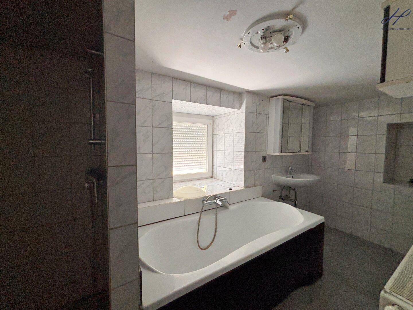 Funktionales Badezimmer mit Badewanne, Duschbereich und Fenster, ausgestattet mit hellen Fliesen.