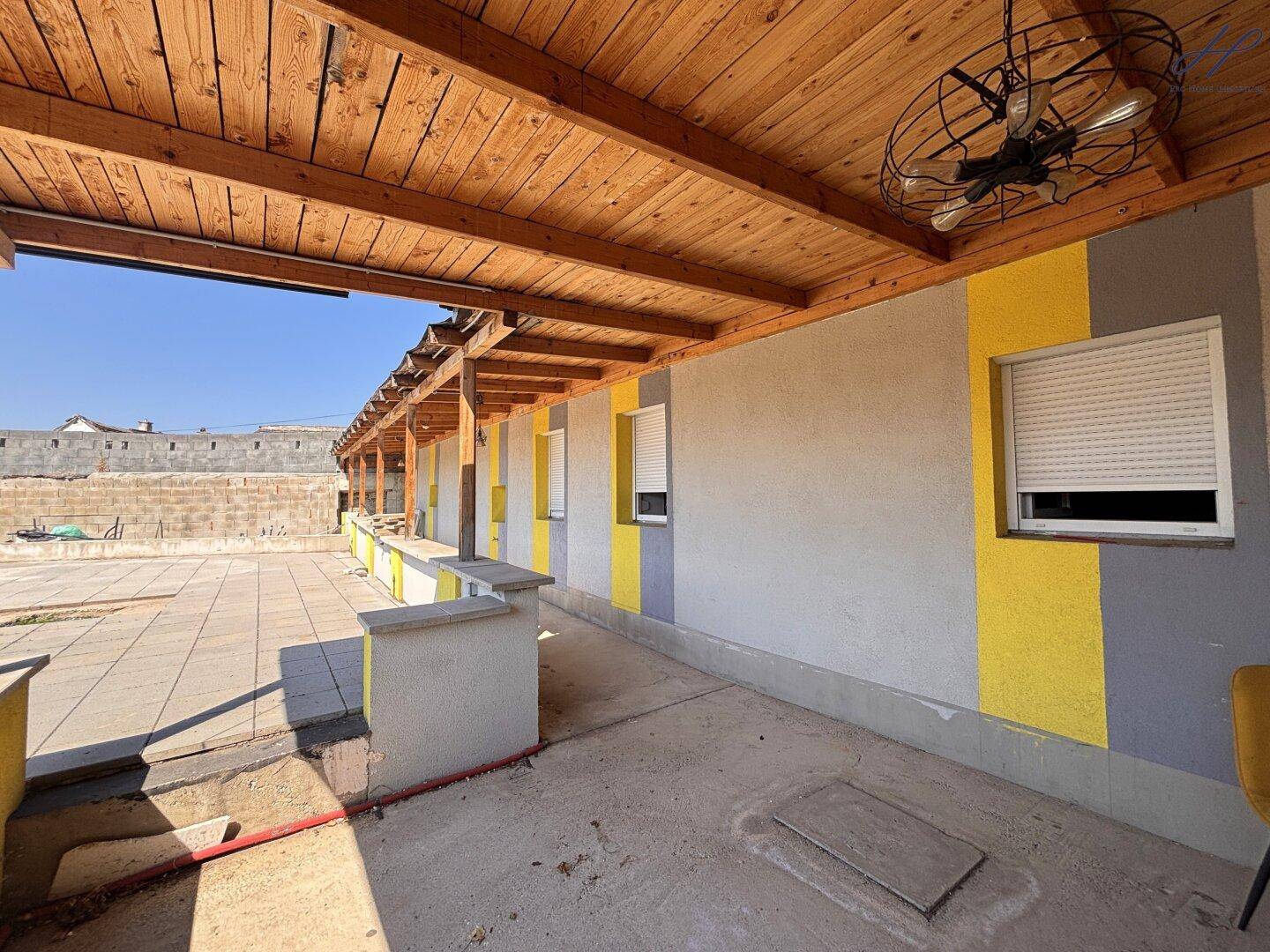 Überdachter Terrassenbereich mit Holzdach, gelb-grauer Fassade und Rollläden an den Fenstern.