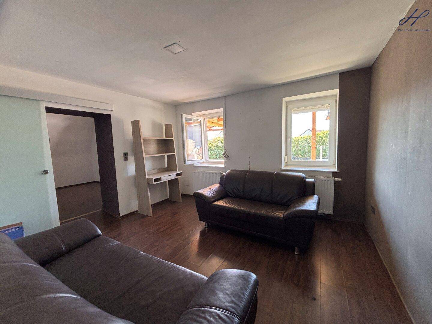 Wohnzimmer mit dunklem Laminatboden, zwei Ledersofas und Fenstern mit Blick ins Grüne.