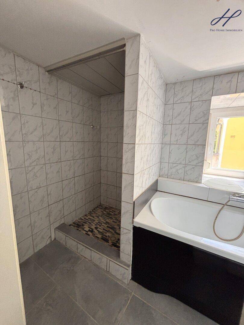 Gefliestes Badezimmer mit integriertem Duschbereich und Badewanne unter einem Fenster.