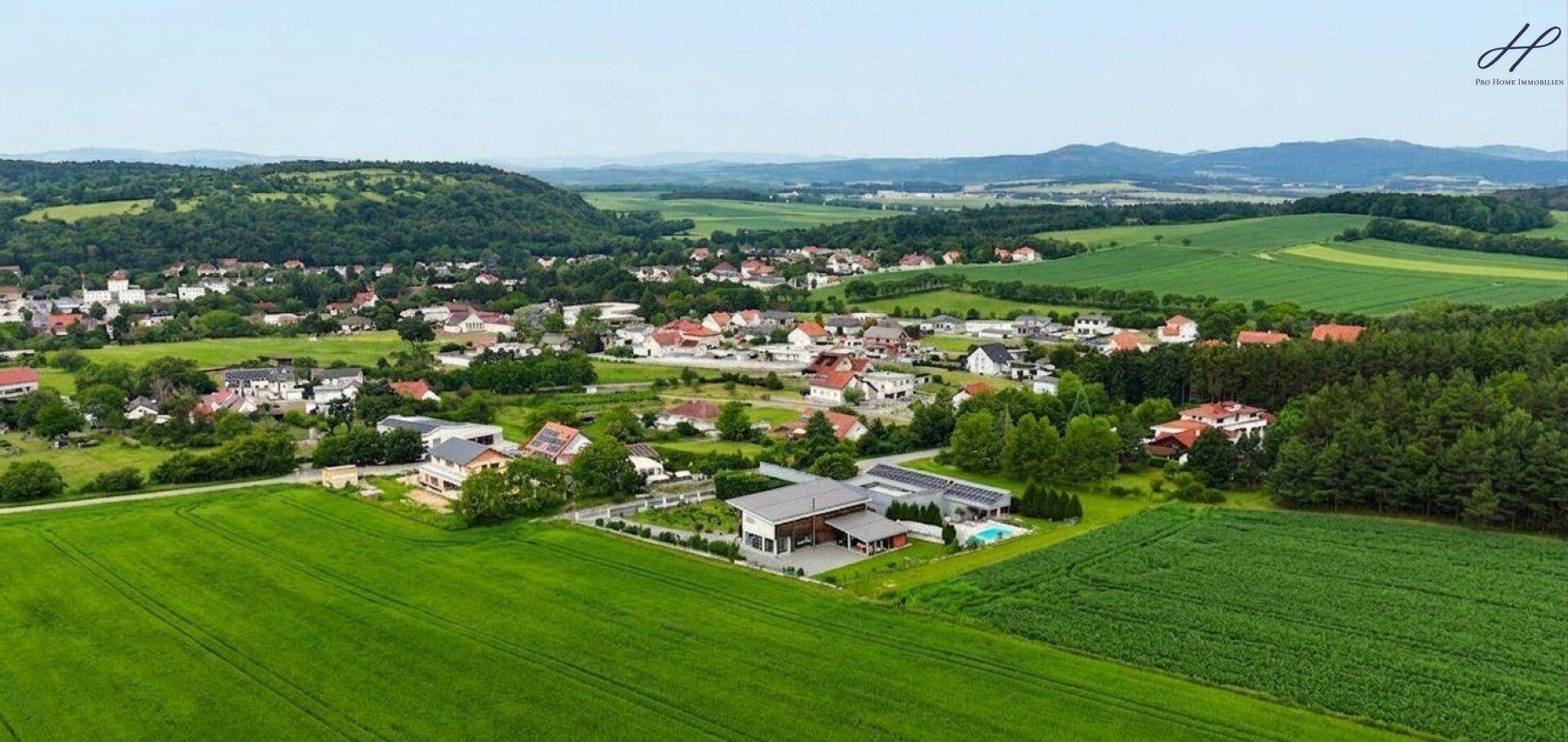 Weitläufige Luftaufnahme eines Dorfes, umgeben von grünen Feldern und Wäldern mit Fernblick.