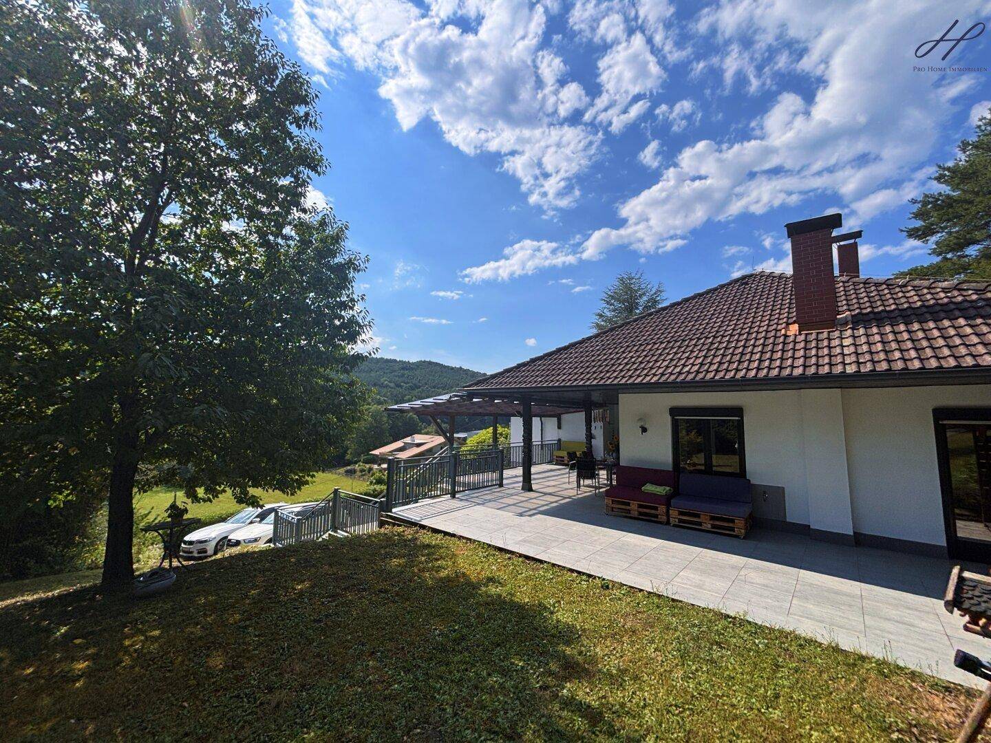 Seitenansicht des Hauses mit weitläufigem Rasen und überdachter Terrasse unter blauem Himmel.