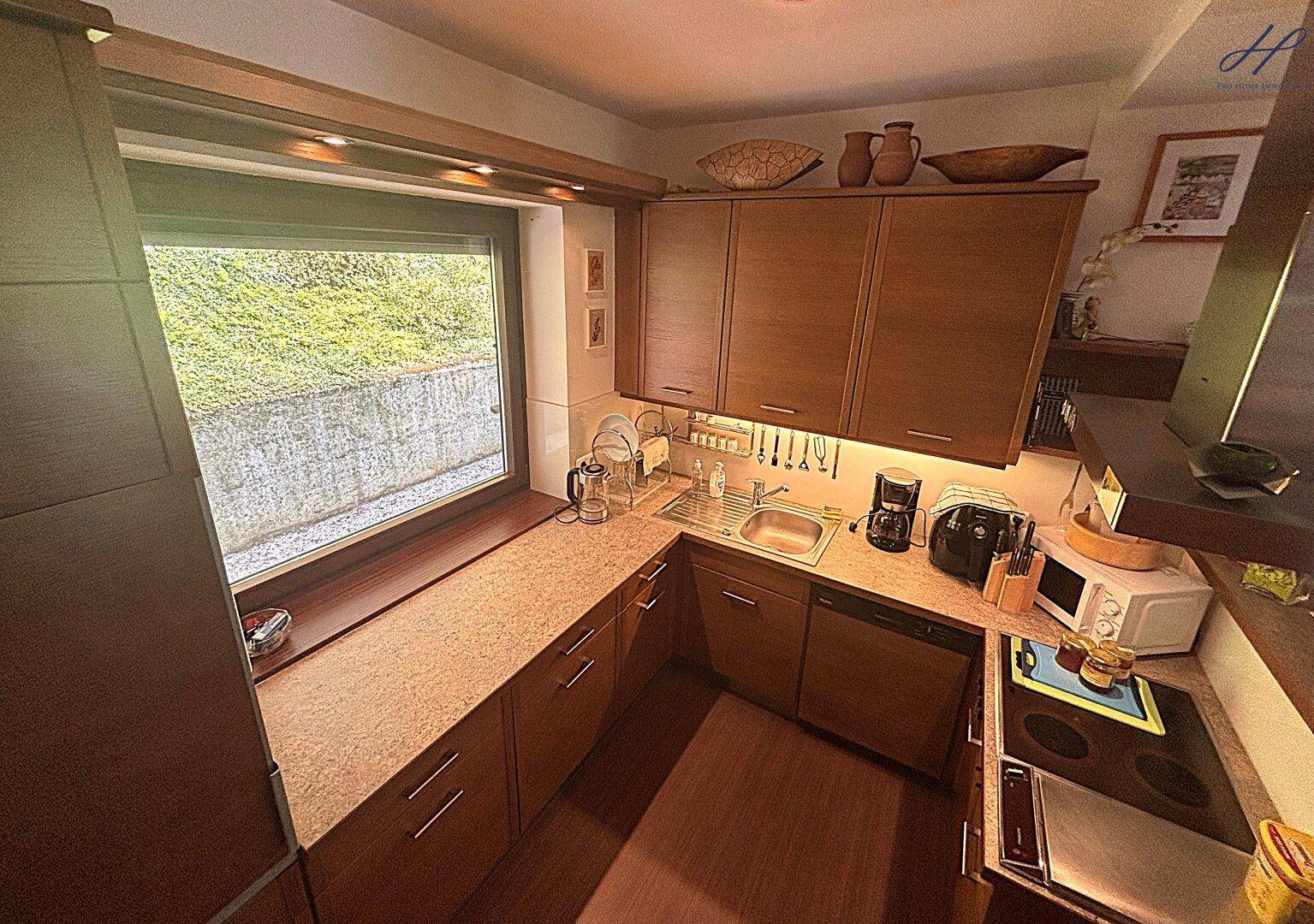 Funktionale Küche mit Holzschränken, Arbeitsplatte und Fenster mit Blick ins Grüne.