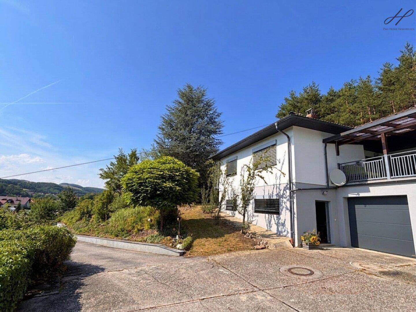 Vorderansicht des Hauses mit integrierter Garage, Zufahrt und umgebender Bepflanzung.