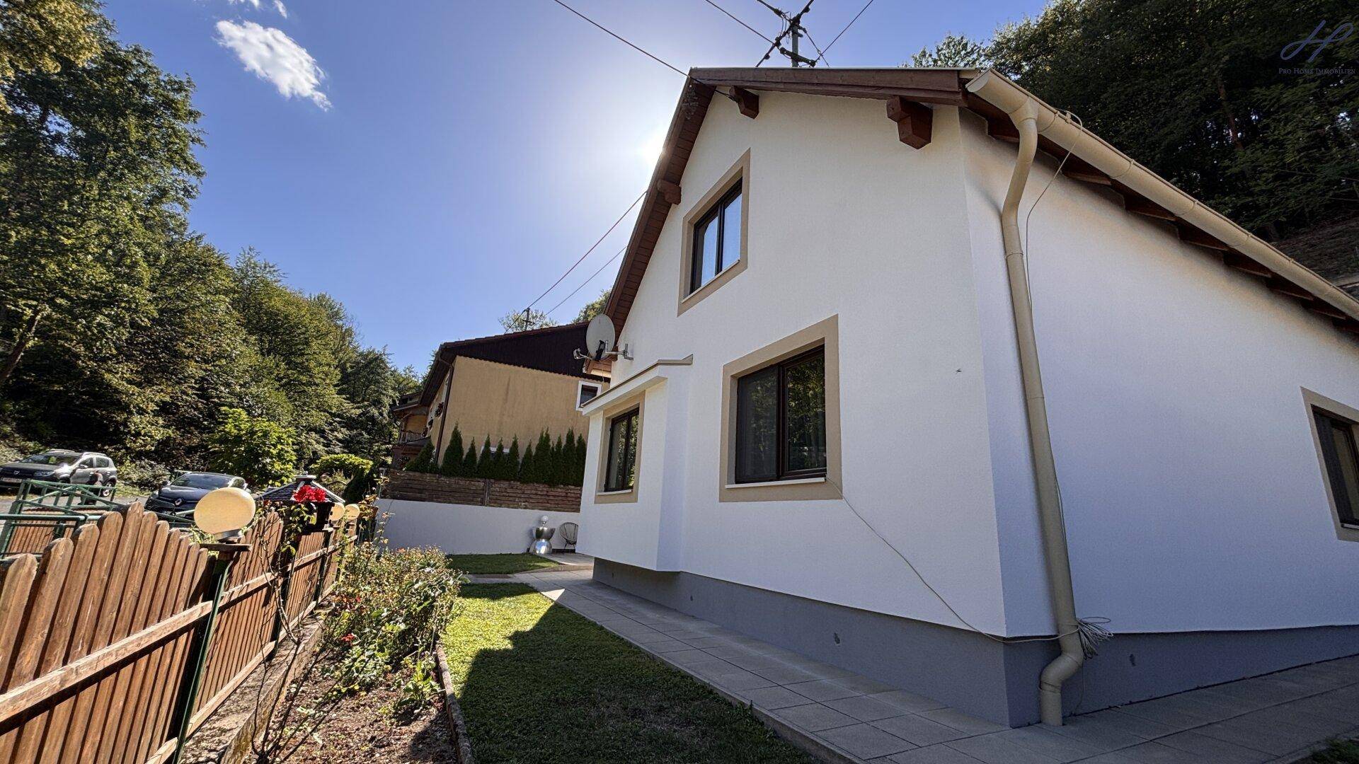 Seitenansicht des Hauses mit weißer Fassade, braunem Dach und sonniger Umgebung.